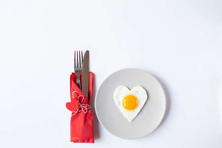 A plate with a fried egg in the form of a heart and cutlery in a red napkin with decorative hearts are on a white background. Valentine's Day breakfast theme. Free space for text.の写真素材