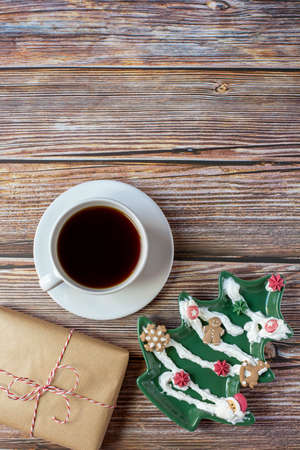 A white cup with black coffee, a Christmas tree-shaped dish decorated with pastry cream and a gift are on a wooden background. Vertical photo. Free space for text.の写真素材