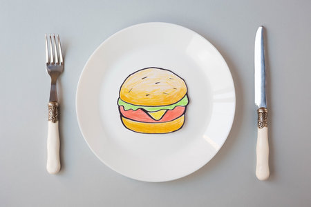 A painted hamburger lies on a white plate next to a knife and fork on a gray background. The topic of sanction and the closure of fast food restaurants. View from the top point.の写真素材