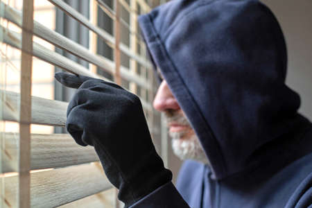 A man in a hooded sweatshirt and black gloves looks out the window through the blinds. The theme of theft and criminology.の写真素材