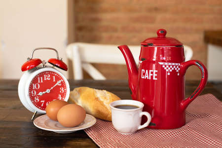 A red coffee pot, a red alarm clock, a white cup of coffee and breakfast are on the wooden table....
