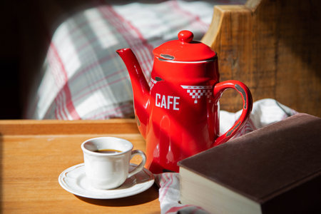 The red coffee pot and black coffee cup are on the bedside table lit with bright light in the morning. Vintage or English style.の写真素材