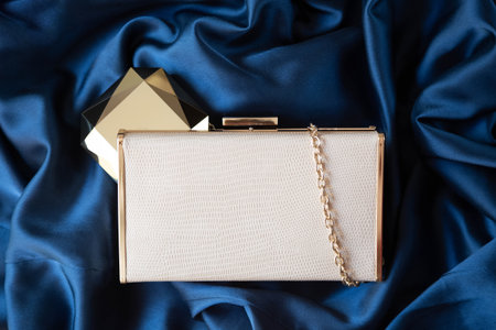 The small beige handbag and gold powder case are set against a dark blue satin material. copyspace.