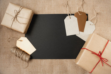 A blank sheet of black paper is surrounded by paper-wrapped gifts and gift tags on a canvas background. Copy space. The theme of decoration of Christmas gifts and seasonal sale.の写真素材