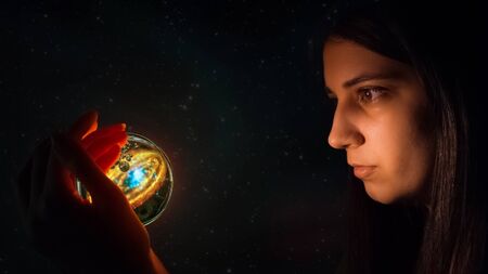 Fortune teller predicts future fate. The Gypsy holds a magic ball in her hand and wonders. Christmas divination. Esoteric and spiritualistic seance. The girl's face lights up the magic ball of fate.の写真素材