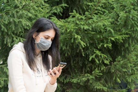 The girl in the medical mask looks at the phone. A woman with long black hair holds a mobile phone....