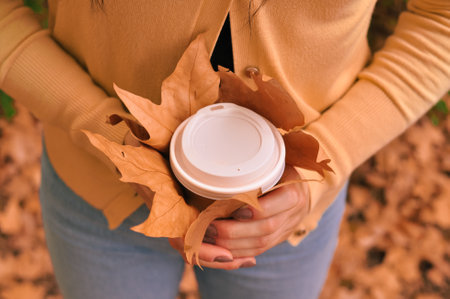 Paper glass of coffee in hand on the orange autumn leaves background . High quality photoの写真素材