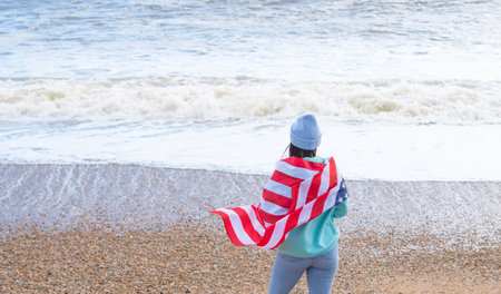 brunette woman in blue sweater and hat with american national flag near sea on the coastlineの写真素材