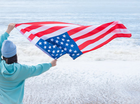 brunette woman in blue sweater and hat with american national flag near sea on the coastlineの写真素材
