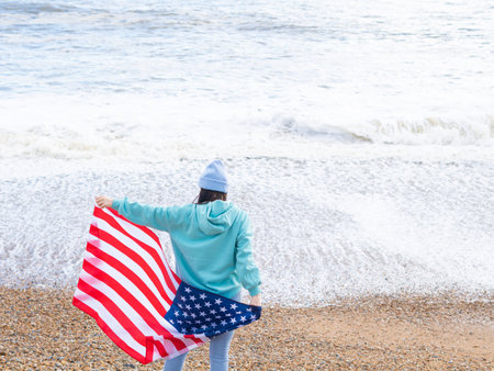 brunette woman in blue sweater and hat with american national flag near sea on the coastlineの写真素材