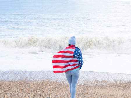 brunette woman in blue sweater and hat with american national flag near sea on the coastlineの写真素材