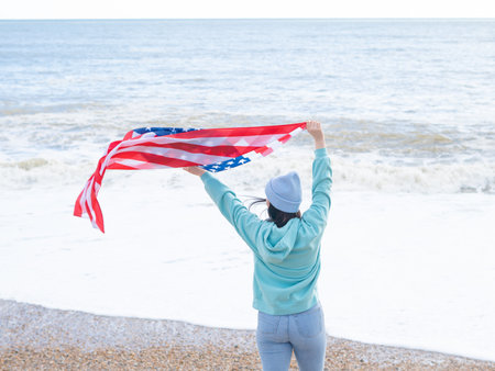 brunette woman in blue sweater and hat with american national flag near sea on the coastlineの写真素材