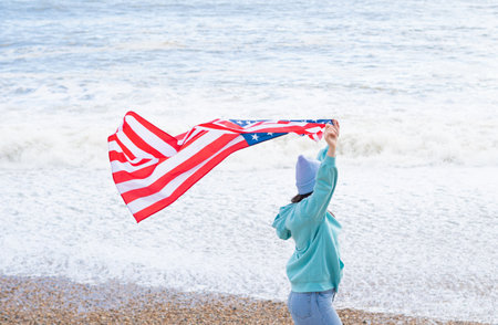 brunette woman in blue sweater and hat with american national flag near sea on the coastlineの写真素材