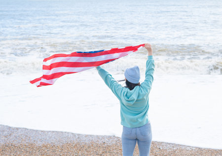 brunette woman in blue sweater and hat with american national flag near sea on the coastlineの写真素材