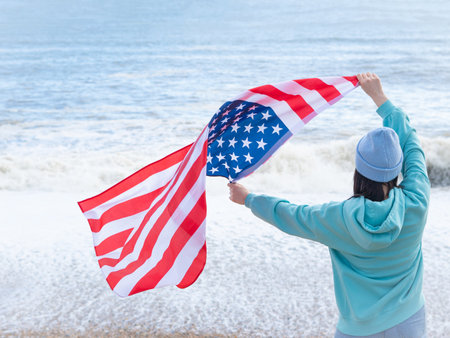 brunette woman in blue sweater and hat with american national flag near sea on the coastlineの写真素材