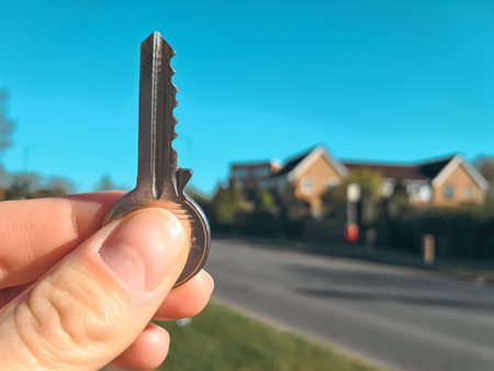 A hand holding a key against a house background, symbolizing real estate dealsの写真素材