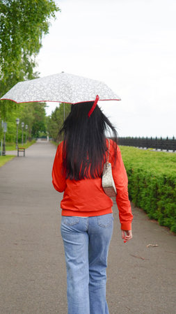 Woman with long hair walks under umbrella in quiet green park after rain.の写真素材