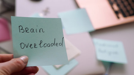 Close-up of a hand holding a sticky note that reads 'Brain Overloaded' on a desk with office items