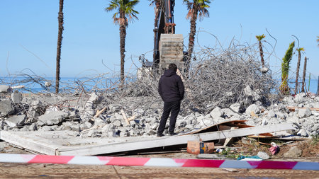 Individual in black clothing surveys rubble at a construction site with palm trees nearby