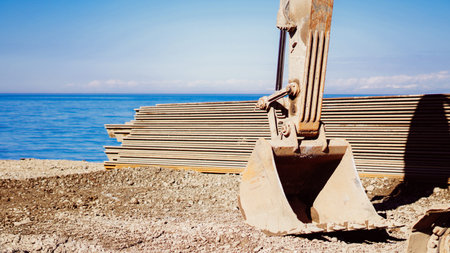Excavator bucket positioned on a construction site by the water with metal sheets stacked nearby