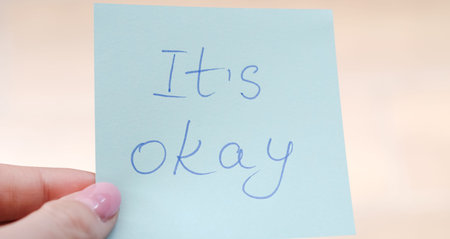 Close-up of a hand holding a blue sticky note with the words "It's okay" written in blue inkの写真素材