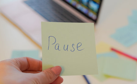Close-up of a hand holding a sticky note that says 'Pause' in a modern office environment