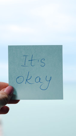 Person hand displays a blue sticky note with the words It's okay against a soft-focus background