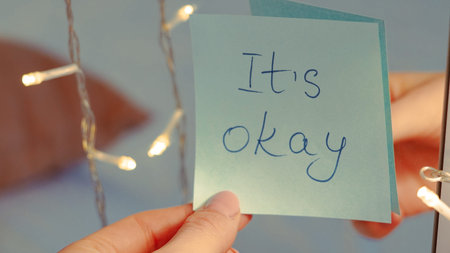 Person's hand displays a sticky note with the words It's okay near glowing string lights