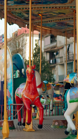 Vibrant red and white carousel horses stand on a ride in an amusement park with buildings nearbyの写真素材