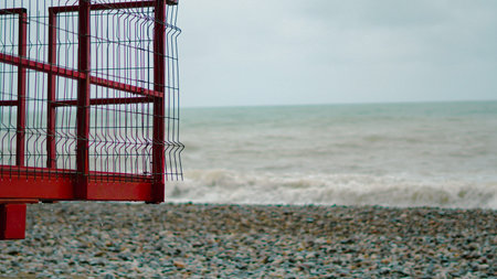 Red lifeguard tower with metal fencing near rocky beach and ocean waves in the backgroundの写真素材