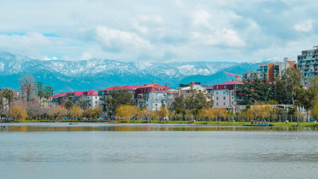 Calm lake reflecting urban buildings and snow-capped mountains under a cloudy sky