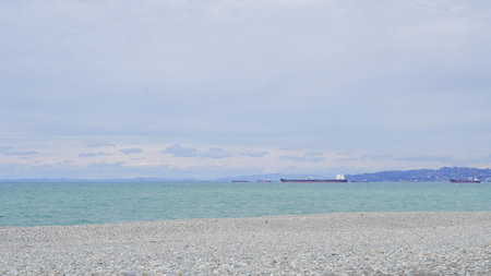 Pebbled beach with calm turquoise water and cargo ships visible in the distance