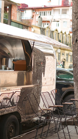 Food truck with shiny metal surface parked beside empty chairs on a bustling city street