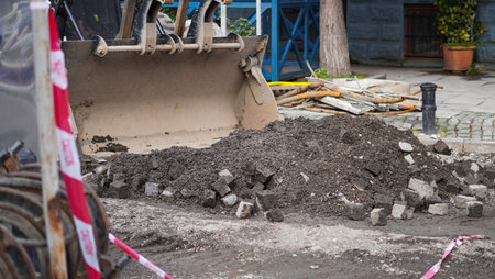 Excavator bucket positioned above a pile of dirt and stones on a city street with barriers