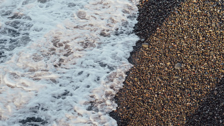 Ocean waves wash over a rocky beach with pebbles and foam along the shoreline