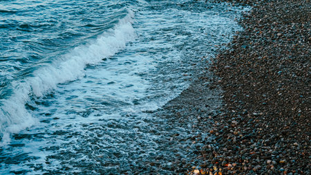 Waves wash over a rocky beach shore with smooth pebbles and clear water visible