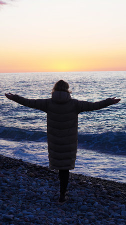 Female figure in a puffer coat stands on a rocky beach with arms open towards the ocean at sunset