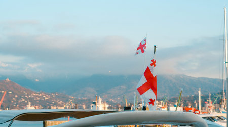 Georgian flags flutter on boats in a harbor with mountains and cloudy sky visible