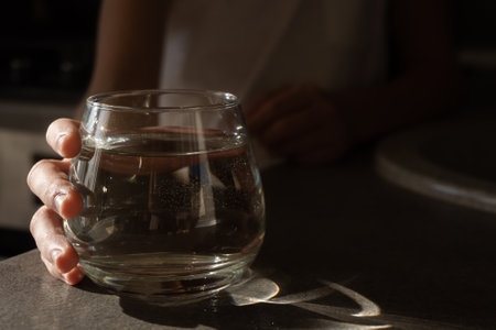 A glass of clean water in a child's hand in daylight with shadows and highlights on the tableの写真素材