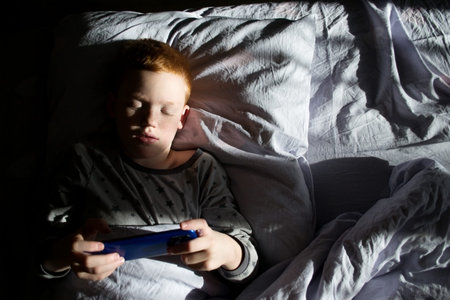 A boy uses his phone while lying in bed in the morning sun.