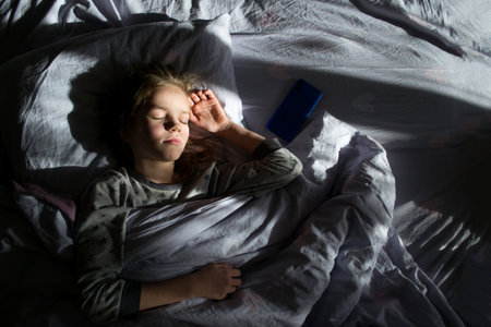 A girl with a phone is resting in bed. A child sleeps on a pillow under a blanket, a phone lies next to himの写真素材