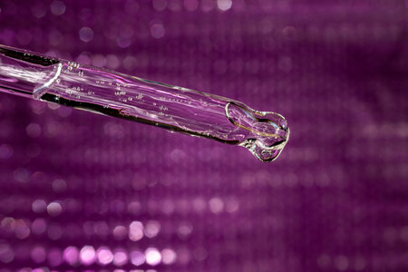 Glass pipette with facial serum on violet background. Essential oil close-up in a pipette. A skin care product with hyaluronic acid in a pipette.の写真素材