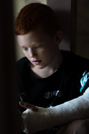 Sad boy with broken arm in plaster bored looking through messages on phoneの写真素材