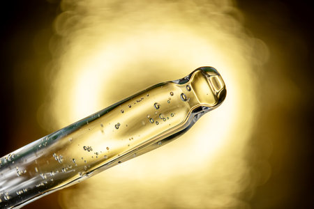Glass pipette with facial serum on gold background. Essential oil close-up in a pipette. A skin care product with hyaluronic acid in a pipette.の写真素材