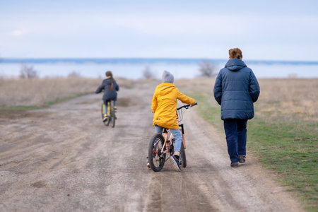 Two sisters are riding bicycles on a cloudy spring or autumn day in nature along a dirt road, their grandmother is walking next to themの写真素材