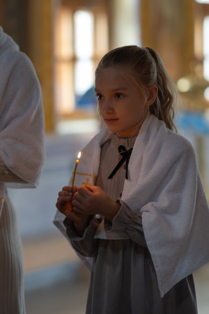 A girl in a church with a white towel over her shoulders holds a burning candle during a service or baptism ceremony. High quality photoの写真素材