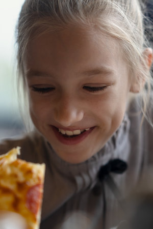 A close-up portrait of a girl biting into a pizza while holding it in her hand. A 9-year-old child with blonde hair eats pizza. High quality photoの写真素材