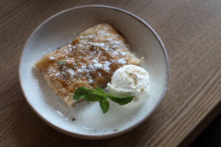 On a white plate lies a slice of golden apple pie or strudel, garnished with a scoop of vanilla ice...