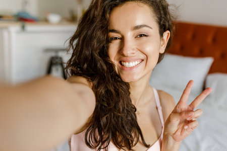 POV of happy Caucasian young brunette woman posing to smartphone camera taking selfie photo and smiling. Joyful positive beautiful lady takes picture of herself at homeの写真素材