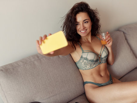 Close up of Caucasian happy female posing to cellphone camera taking pictures posing in underwear at home. Joyful pretty woman in sexy lingerie takes selfie photo on smartphoneの写真素材